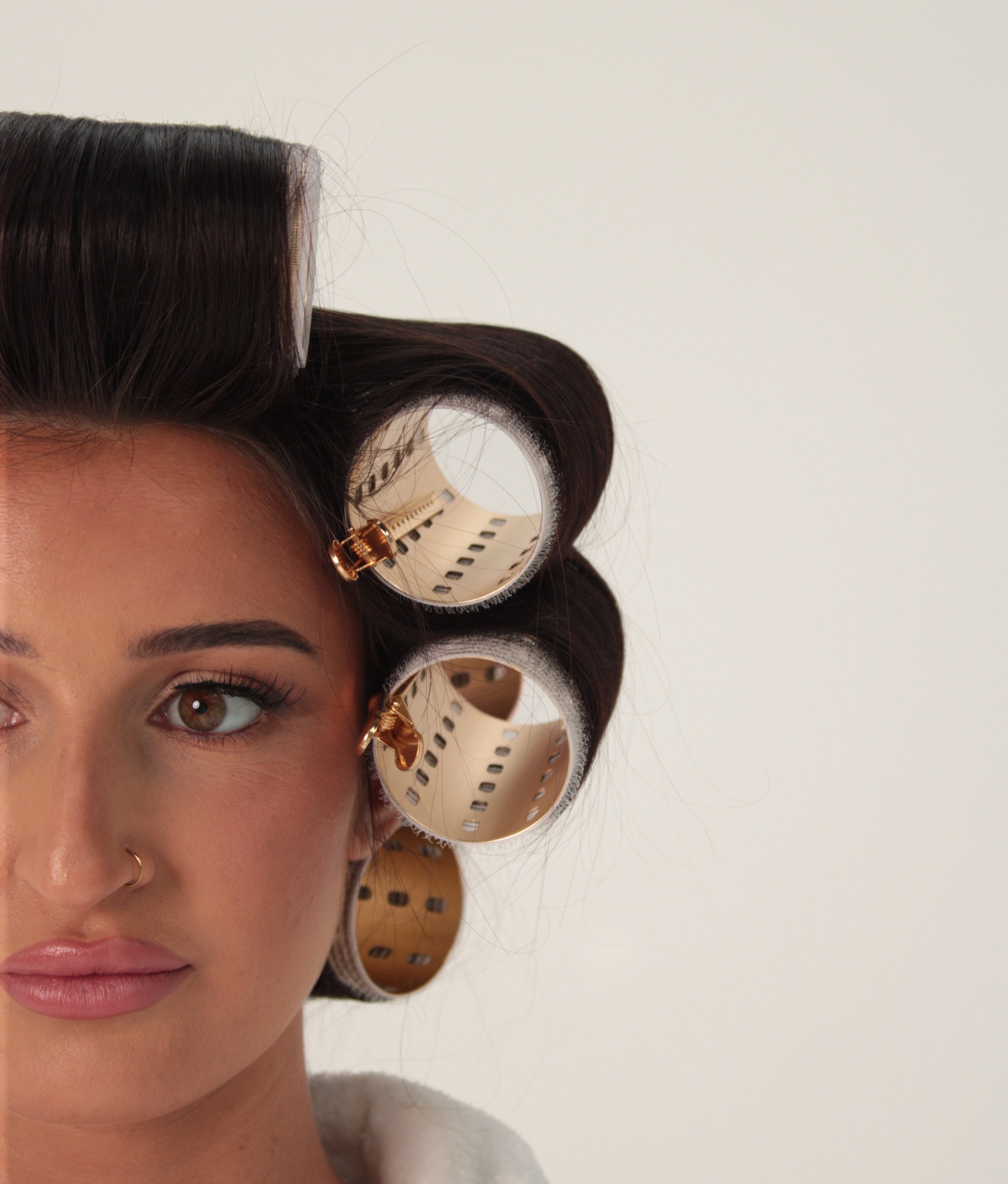 A close-up of a woman with dark hair styled in large curlers, showing the beige and gold plastic curling devices against a plain light background.