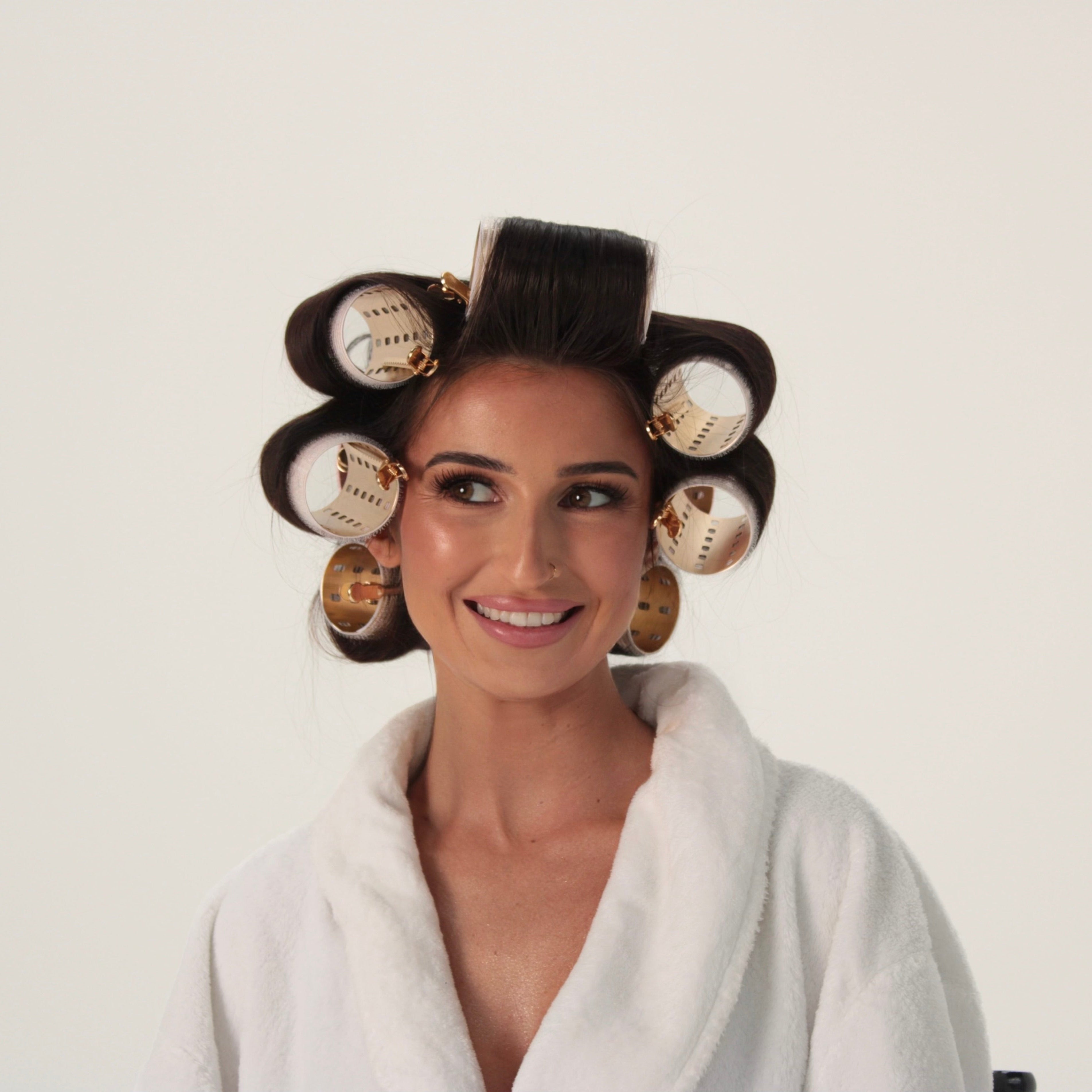 A woman with dark hair styled in large eight gold velcro rollers wearing a white bathrobe, photographed against a plain light background.