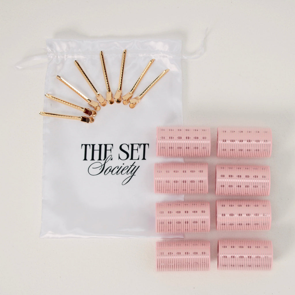 A hair styling set from The Set Society on a white background, featuring eight pink barrel curlers and eight gold metal hair pins inside a clear drawstring pouch.