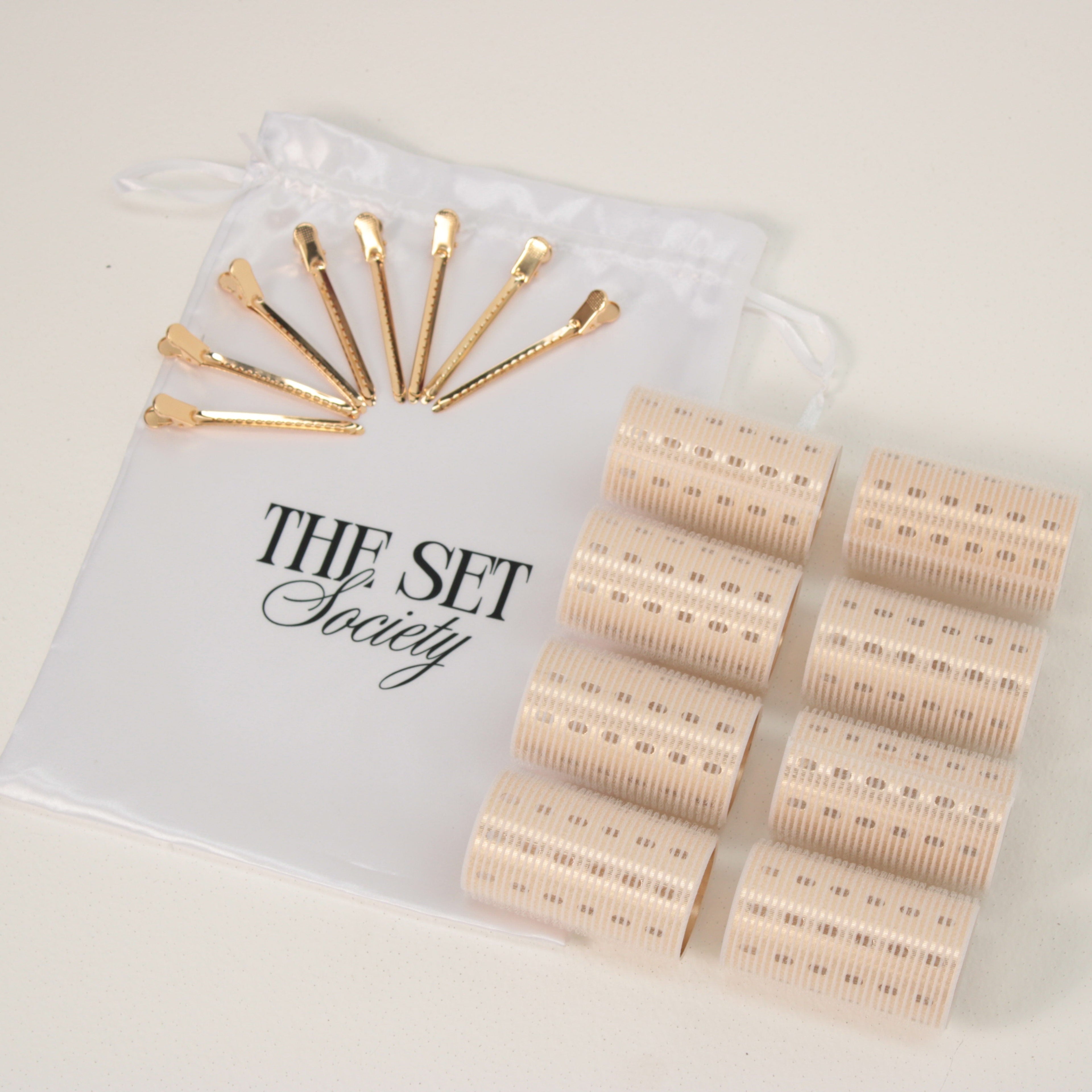 A hair styling set from The Set Society, featuring eight gold metal hair clips arranged on a white drawstring pouch, alongside multiple beige textured hair rollers, photographed on a clean off-white background.