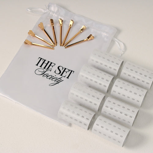 A white sheer drawstring pouch printed with 'THE SET Society' and gold hair clips, shown next to eight white velcro hair rollers
